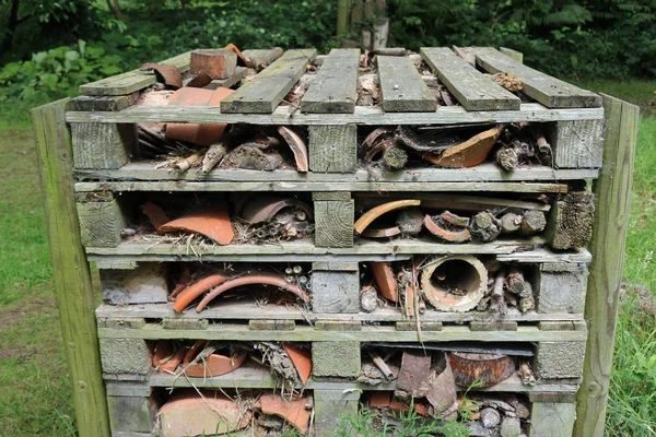 Bug hotel in a frame of wooden pallets containing logs, branches, bamboo stems and broken clay flower pots surrounded by grass with trees in the background.