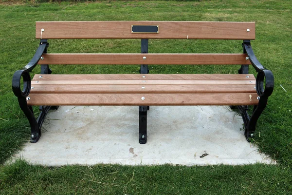 Metal framed wooden seat with a plaque standing on a concrete plinth surrounded by a grass lawn.