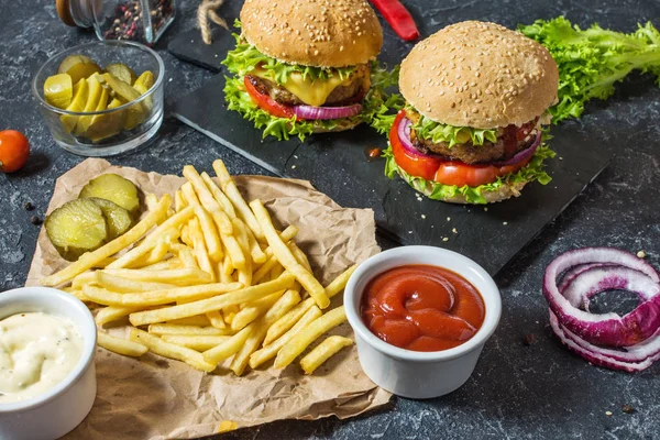 Two beef burgers, fries potatoes and cucumber on dark stone table.