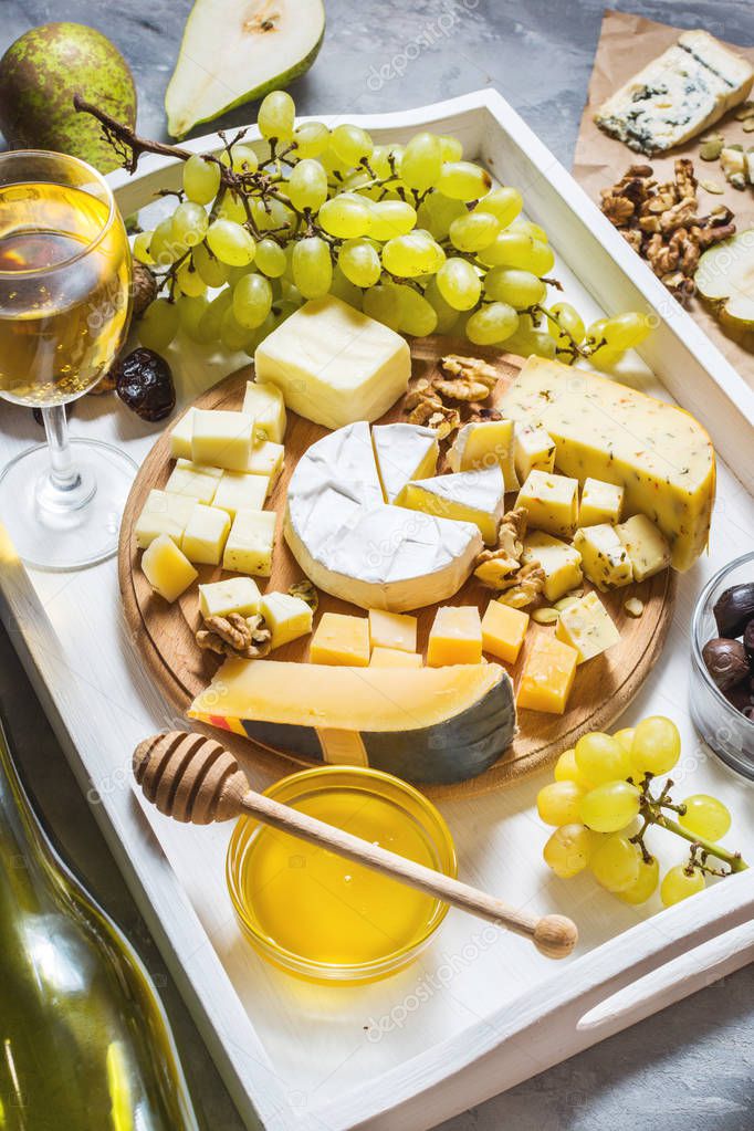 Different types of cheese on wooden board, olive, fruits, almond and wine glasses on white tray on concrete table