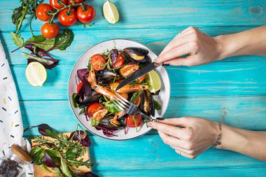 Taze deniz ürünleri salatası, midye, karides, taze sebze ve otlar mavi ahşap masa üzerinde. Eller çatal ve bıçak tutun. Üstten Görünüm