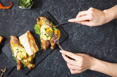 Kadın eli Benedict yumurtalı sandviç, siyah taşlı pastırma ve salata tutuyor. Üst görünüm