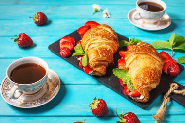 Lezzetli bir kahvaltı. Çilekli taze kruvasanlar ve mavi ahşap arka planda siyah kahve.