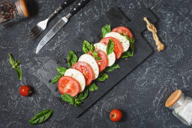 Geleneksel İtalyan Caprese Salatası - dilimlenmiş domates, mozzarella peyniri ve koyu taş arka planda fesleğen, üst manzara