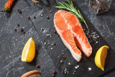 Siyah taş arka planda tuz, biber, limon ve biberiyeli taze somon bifteği.