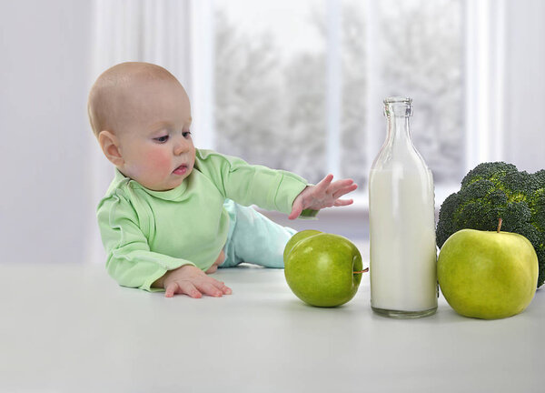 Маленький ребенок тянется за свежим зеленым яблоком на белом столе
