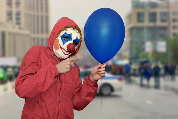 Person in a clown mask scares on the street with a balloon and a needle. Festive fun. Malice concept