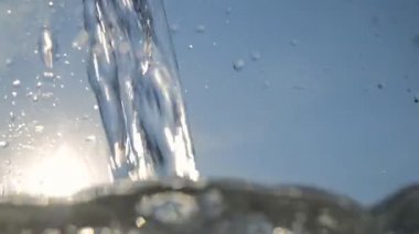 Mavi gökyüzüne ve parlak güneşe karşı şeffaf bir kabı temiz suyla doldurmak. Cam bir yüzeye sıçrama ve damlalarla. 4K video, yakın çekim. Soyut.