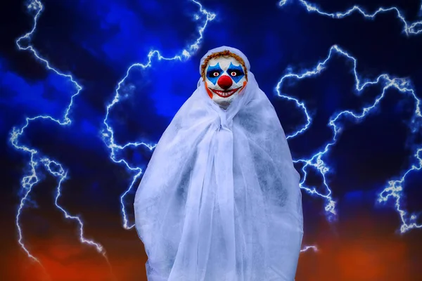 Evil clown in a mask stands against a blue night sky with lightning. Wrapped in white cloth. Disguised and scary person. Halloween holiday.