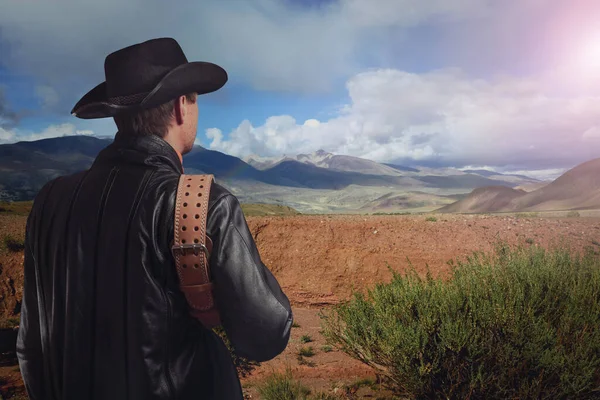 Kovboy dağlardaki mesafeye bakıyor. Deri ceketli ve kovboy şapkalı bir adam kemer tutuyor. Batı kültürü.