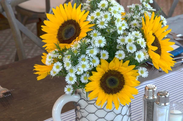 Fresh yellow sunflowers and white daisies bloom in a white vase covered with chicken wire on a rustic farm table.