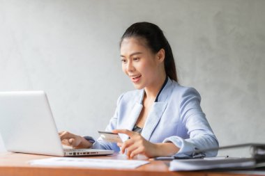 Genç Asyalı bir kadın internet üzerinden alışveriş yapmak için bilgisayarını ve kredi kartını kullanıyor, gülümsüyor ve iş kadını yaşam tarzı kavramını mutlu ediyor.