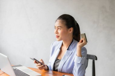 Genç Asyalı bir kadın internet üzerinden alışveriş yapmak için bilgisayarını ve kredi kartını kullanıyor, gülümsüyor ve iş kadını yaşam tarzı kavramını mutlu ediyor.