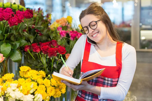 Female attractive florist accepting orders over a phone and taking notes in a notebook in a flower shop. Woman assistant or owner in floral design studio, making decorations. Flowers delivery service.