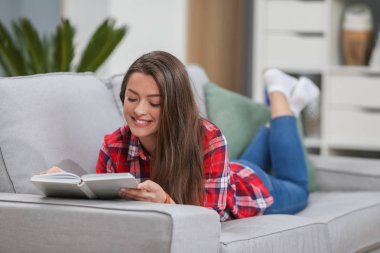 Rahat kıyafetler bir kitap okuma ve bir kanepe odasında yatarken gülümseyen güzel kadın.