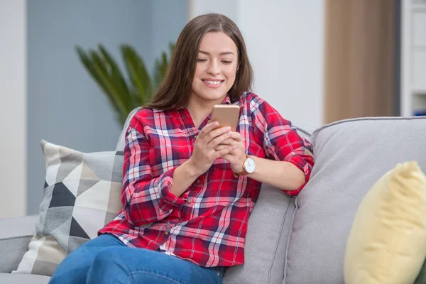 Good-looking, magnificent, exquisite, delicate, winsome, lovable woman sit on cozy couch in shirt and denim jeans in modern light interior and look at phone screen make hollywood smile