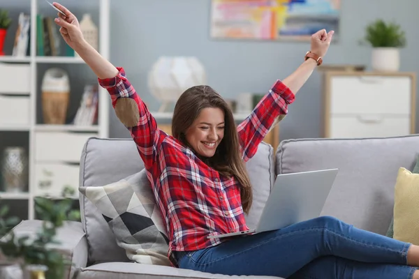Kredi kartı tutan ve dizüstü bilgisayar kullanan güzel kadın. Online alışveriş kavramı. Kız evde kanepede rahatlatıcı