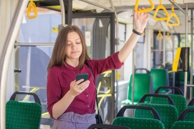 Modern tramvayda hareket ederken akıllı telefonla ayakta duran genç kadın yolcu, toplu taşıma araçlarında yolculuğun keyfini çıkarıyor