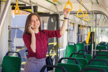 Modern tramvayda hareket ederken akıllı telefonla ayakta duran genç kadın yolcu, toplu taşıma araçlarında yolculuğun keyfini çıkarıyor