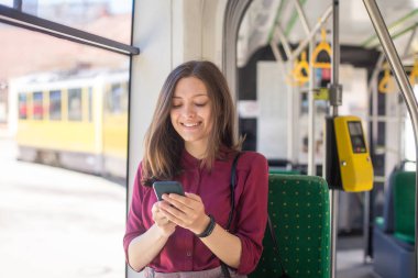 Modern tramvayda hareket ederken akıllı telefonla ayakta duran genç kadın yolcu, toplu taşıma araçlarında yolculuğun keyfini çıkarıyor
