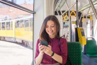 Modern tramvayda hareket ederken akıllı telefonla ayakta duran genç kadın yolcu, toplu taşıma araçlarında yolculuğun keyfini çıkarıyor