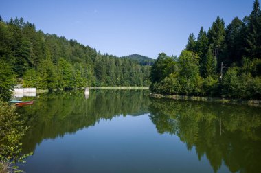 Sakin bir göl, temiz bir gökyüzünün altındaki ormanı yansıtır. Tekneler kıyıda dinleniyor, ziyaretçileri sakin suları keşfetmeye ve doğanın güzelliğinin tadını çıkarmaya davet ediyor..