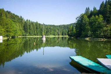 Sakin bir göl çevresindeki ormanı ve gökyüzünü yansıtır. Ön planda bir rıhtım, ziyaretçileri sakin suların ve doğal güzelliğin tadını çıkarmaya davet ediyor..