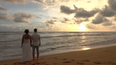 Romantik bir tarihte aşık genç çift, el ele tutuşarak, sahilde gün batımı karşılar