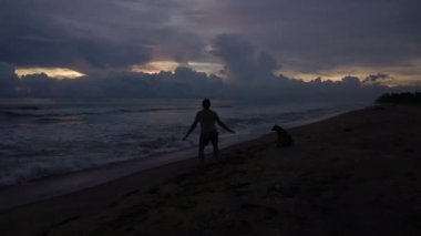 Mutlu genç erkek siluet gün batımında sahilde atlıyor, el kaldırıyor. Yavaş çekim. 240fps hızında çekildi