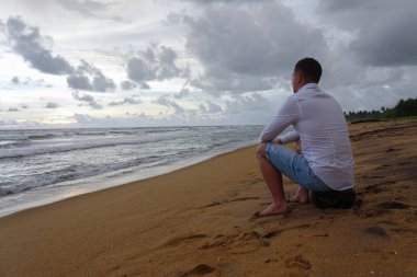 Beyaz gömlekli genç adam tropik bir okyanus sahilinde gün batımıyla buluşuyor