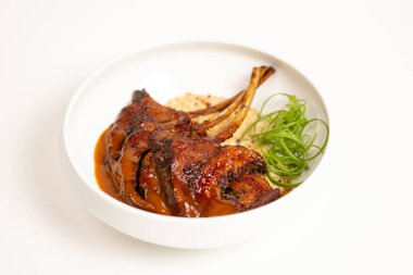 Zengin kahverengi soslu, sulu ızgara kuzu pirzolası, kremalı garnitür ve otlarla servis edilmiş, gıda tasarımı projeleri için beyaz bir arka planda fotoğraflanmış..