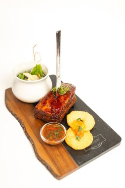 Top view of grilled pork ribs with sauce, bread, and herbs on a wooden board. Stylish presentation on a clean white background.