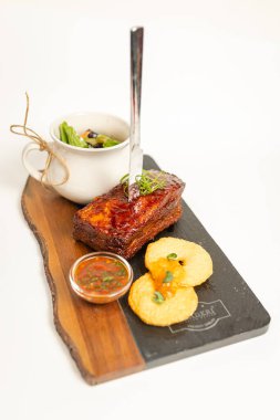 A slice of grilled meat with sauce, bread, and herbs on a stone board. Elegant plating in a restaurant setting against a white background.