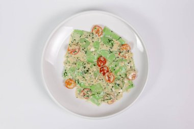 Bitki özlü makarnadan yapılan taze yeşil ravioli kremalı peynir sosu, yarım kiraz domatesleri ve beyaz bir tabakta doğranmış bitkilerle servis ediliyor. Minimalist sunum, stüdyo yemek fotoğrafçılığı.