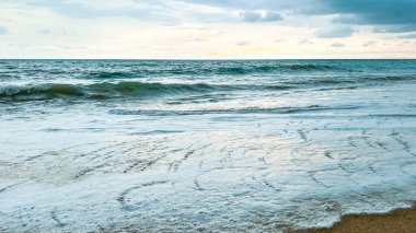 Wide view of ocean waves, foam, and sand. Cloudy sky and calm water create a peaceful, serene atmosphere.