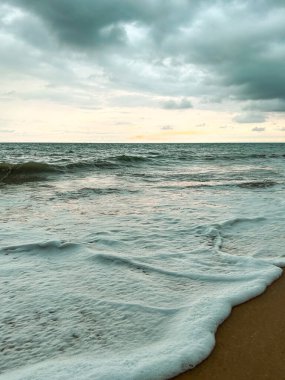Close-up of sea foam washing over sand. Dark clouds and calm ocean create a peaceful, dramatic atmosphere.