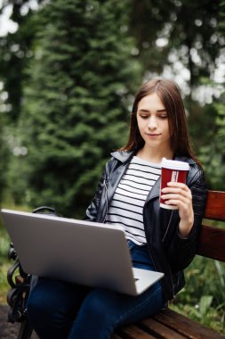 Genç öğrenci dizüstü bilgisayar ile içer bankta oturan ve bir yaz parkta okuma kahve fincanı.