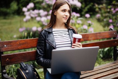 Parkta motive serbest meslek. Kahve içme ve laptop ile online çalışan genç esmer kız bir bankta.