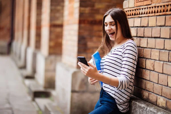 Half length portrait of cheerful young woman standing in the street beside brick wall, sending message on smartphone.