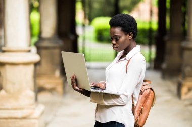 Portrait of smiling African american student doing homework outdoors using laptop for research .Positive dark skinned hipster girl styling in university campus preparing for examinations outdoors clipart