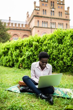 Genç iş kadını afro saç modeli ile bir dizüstü bilgisayar ekranında açık havada çalışırken gülümsüyor. Uluslararası öğrenci wi-fi için bağlı bir taşınabilir bilgisayar tarafından online sınavlar için hazırlanıyor.