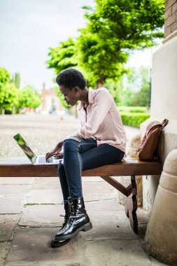 Genç afro Amerikan erkek bu uzaktan bir kamu kullanarak yeni bir proje üzerinde çalışma uzman ağ wifi ahşap bir bankta otururken. Kampüs çalışmak için dizüstü bilgisayar kullanan güzel öğrenci kız