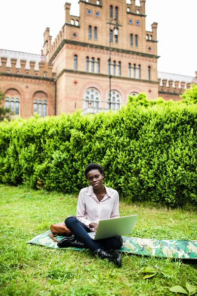 African american international student typing on laptop preparing for examination outdoors in campus enjoying weather. Smiling female copywriter work on publication for network sitting on grass