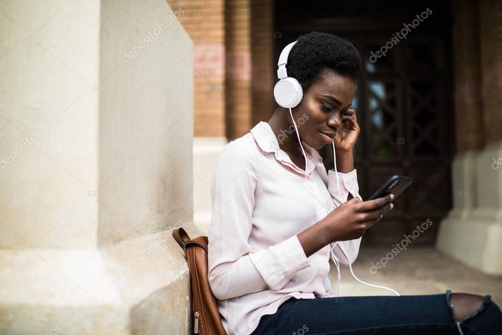 Beautiful afro american girl listening to music through headphones sitting on a bench.