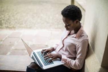 Genç afro Amerikan erkek bu uzaktan bir kamu kullanarak yeni bir proje üzerinde çalışma uzman ağ wifi ahşap bir bankta otururken. Kampüs çalışmak için dizüstü bilgisayar kullanan güzel öğrenci kız