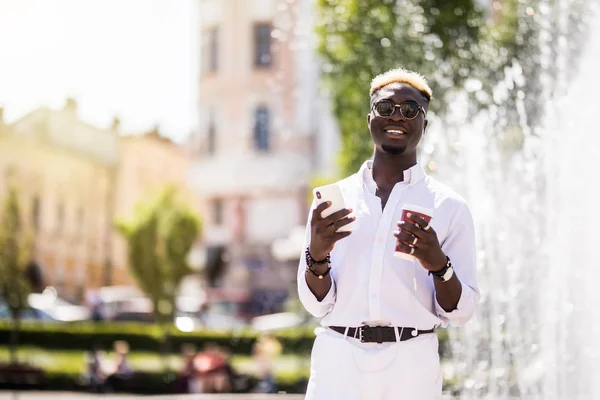 Afro american man setting playlist on smartphone the street. Young Afro American man with earphones using phone and surfing music on internet near city fountains