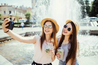 Güzel arkadaşlar modern akıllı telefon kameraya poz, selfie yapmak, birlikte sokak içki sallamak açık oturup, olumlu ifadeler var.