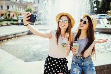 İki neşeli kadın karşı çeşme ile güler yüzlü poz, modern cep telefonunda selfie yapmak veya görüntülü görüşme konuşma online, yurt dışında resort hakkında onların gösterim payı