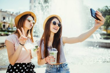 Two happy women in sunglasses and hats are having nice time together in the city taking selfie near fountain on the swuare clipart