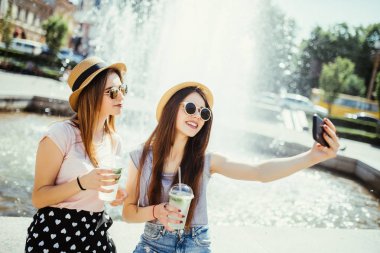 Güneş gözlüğü ve şapka yakınındaki şehir Çeşme'de iki genç kız. Arkadaşlarım kendi fotoğraf yapıyor ve şehirde sokak gülüyor kokteyl içmek .